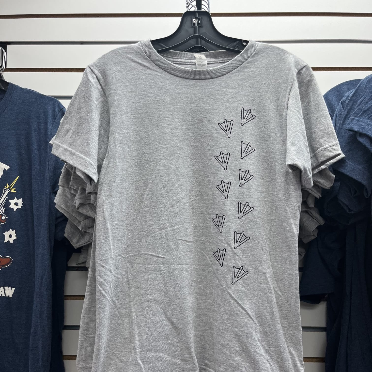 Gray t-shirt with a silly goose foot print design on a hanger against a white wall with other shirts.