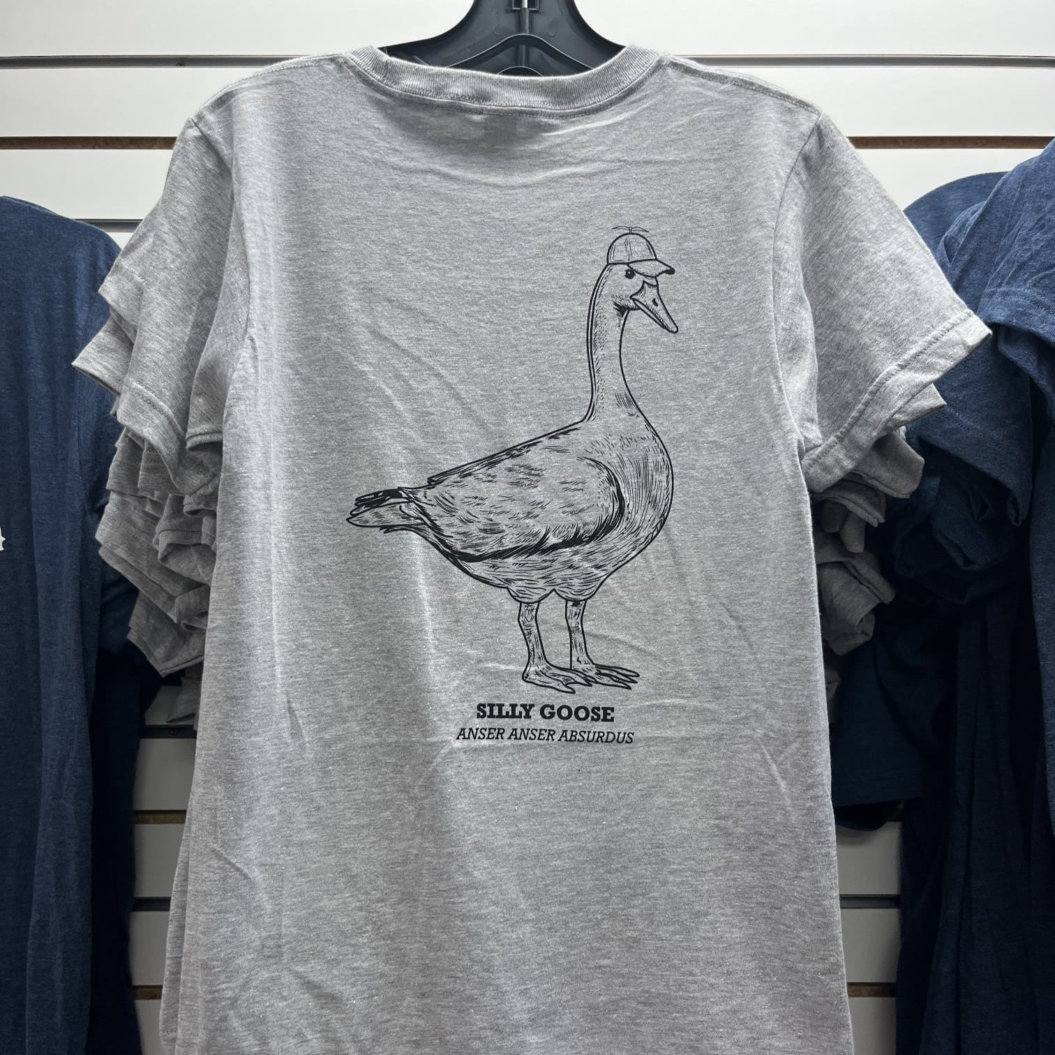 Gray t-shirt with a goose graphic and text on a hanger against a striped wall.