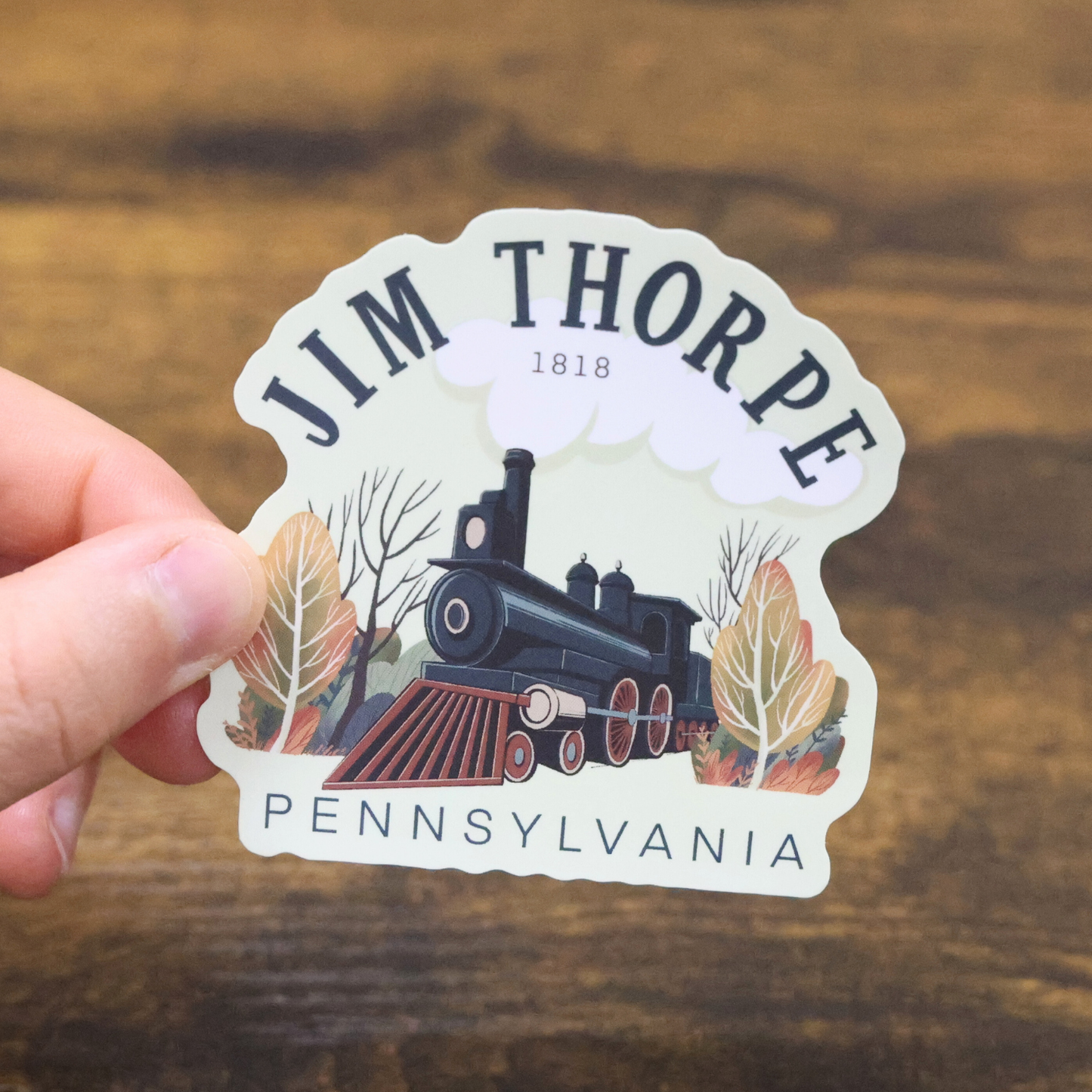 Sticker of a steam train with 'Jim Thorpe, Pennsylvania' text held by a hand on a wooden surface.