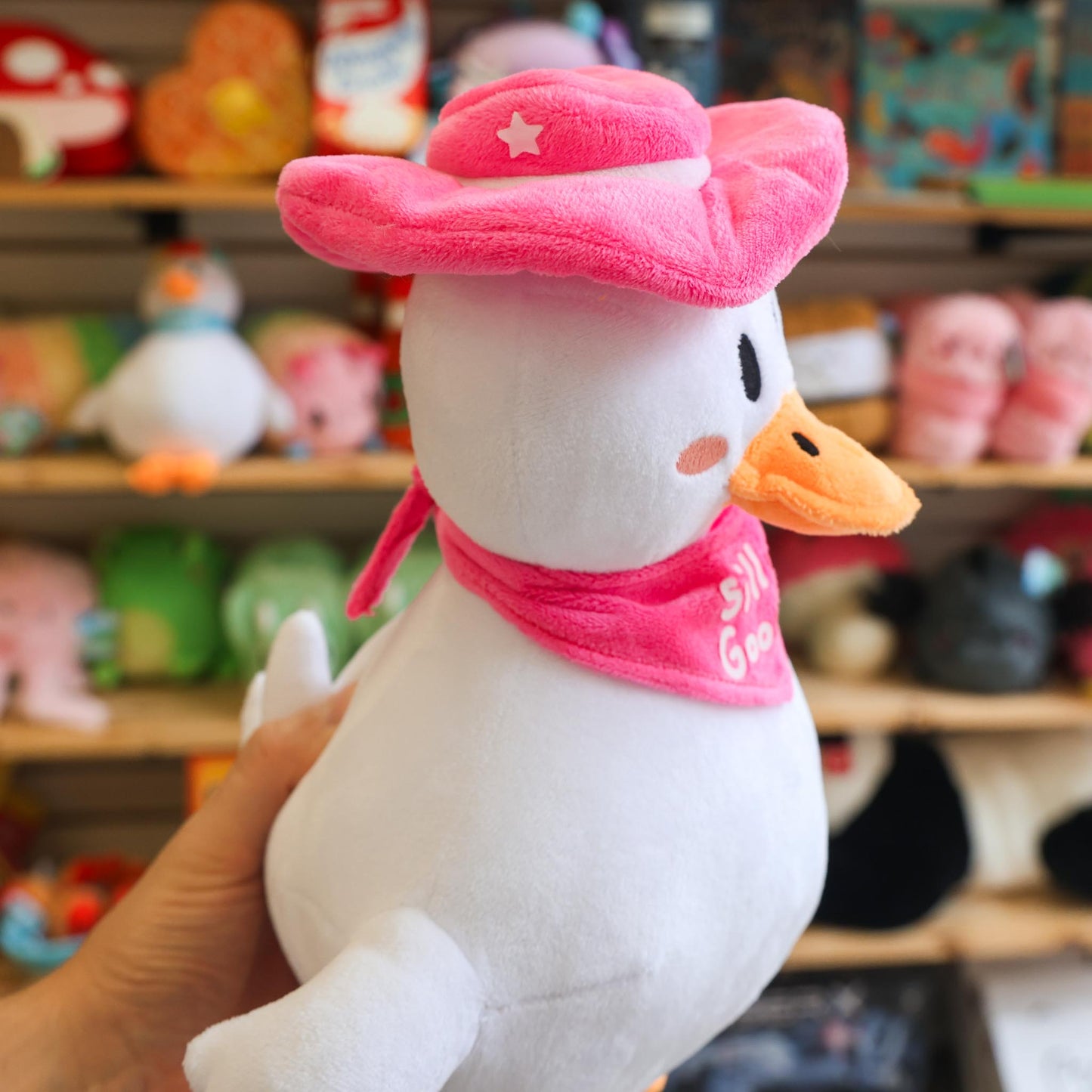Plush toy goose wearing a pink cowboy hat and bandana in front of a store shelf with various toys.