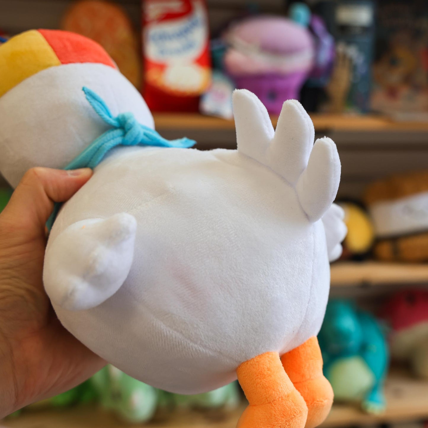 White plush toy with orange feet held by a hand against a blurred background of toys and shelves.
