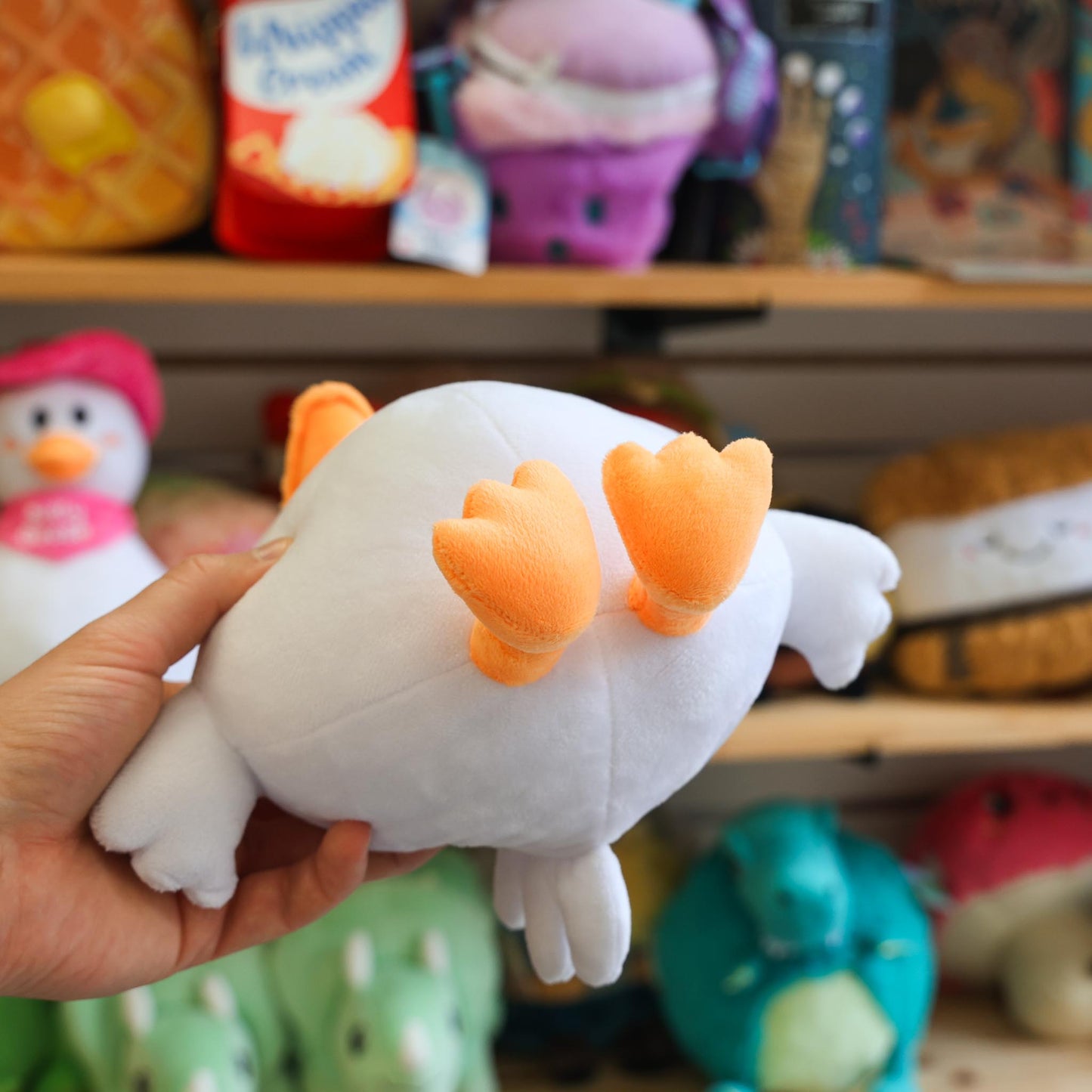 White plush toy with orange accents held in front of a shelf with various toys.