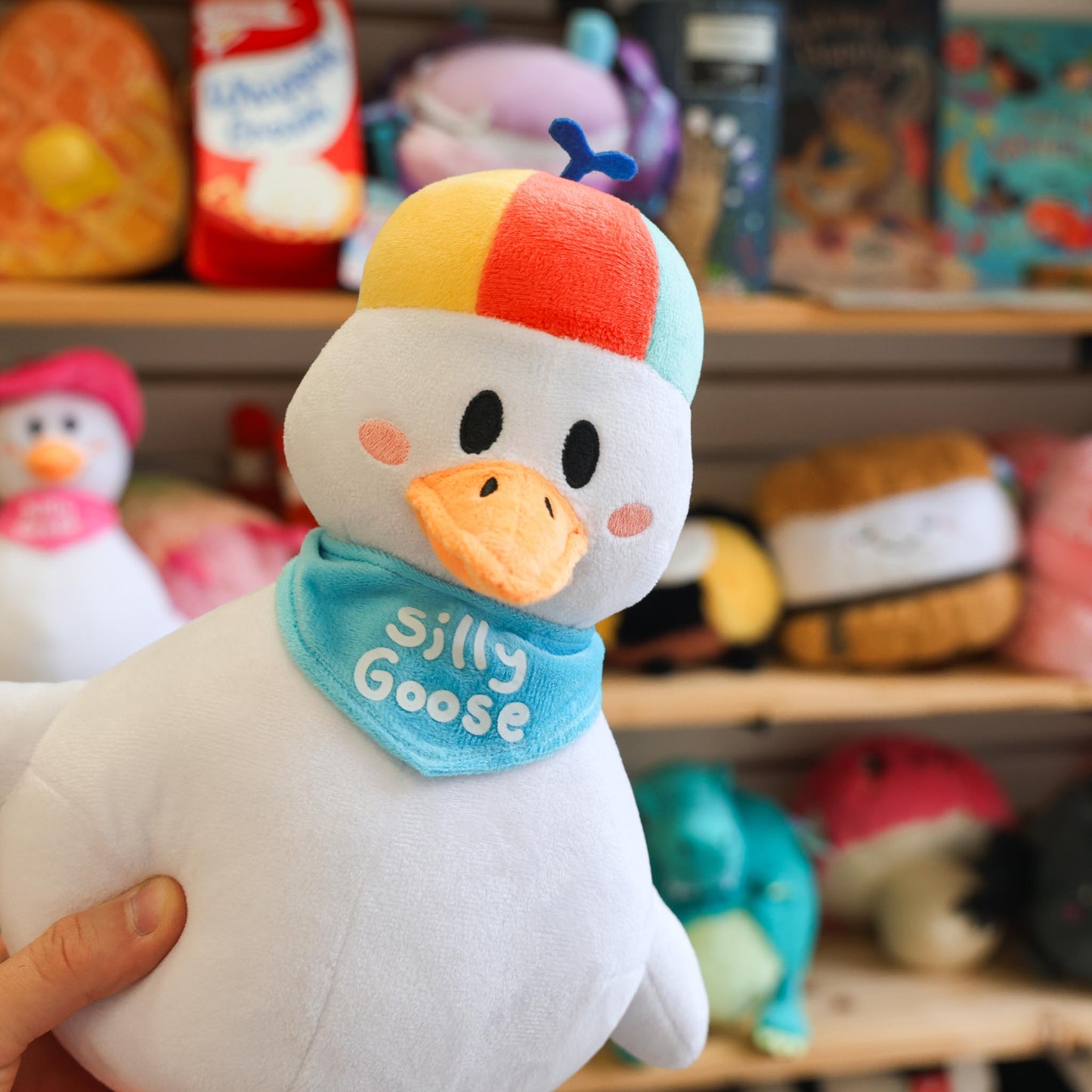 Plush toy goose with colorful hat and 'Silly Goose' bandana held in front of a shelf with other toys.