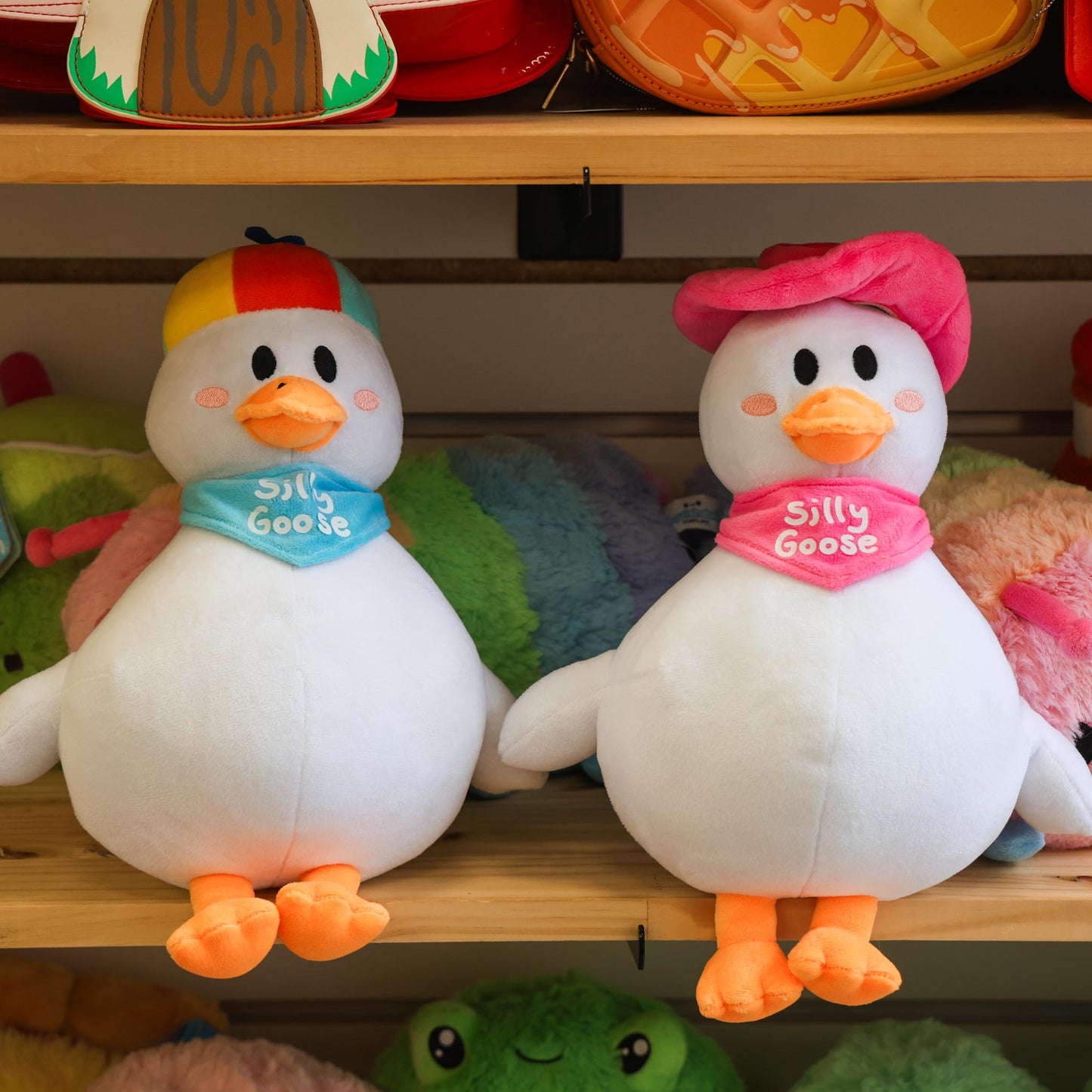 Two plush goose with colorful hats and scarves on a shelf with other toys.