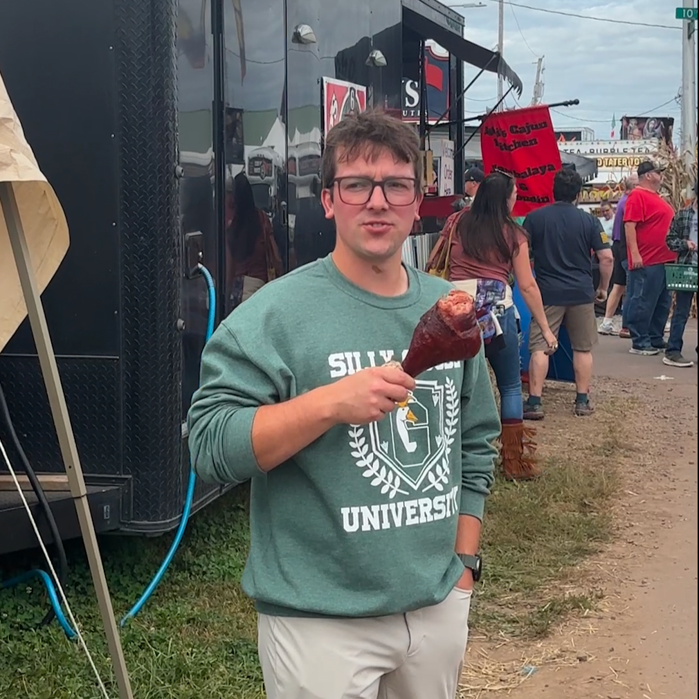 Man holding a giant turkey leg with a silly goose university sweatshirt outdoors near a black trailer with people in the background.