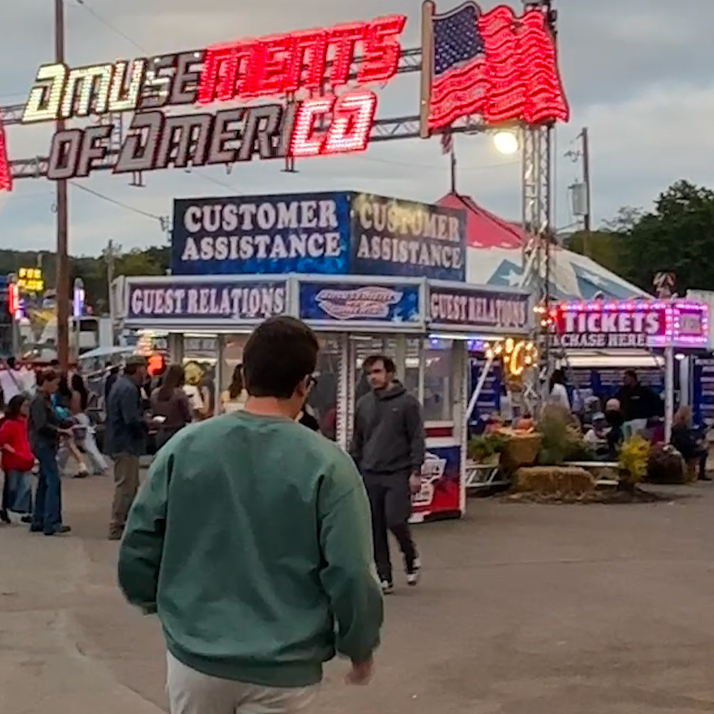 Amusement park entrance with a large sign and people walking around. Back of silly goose university crewneck