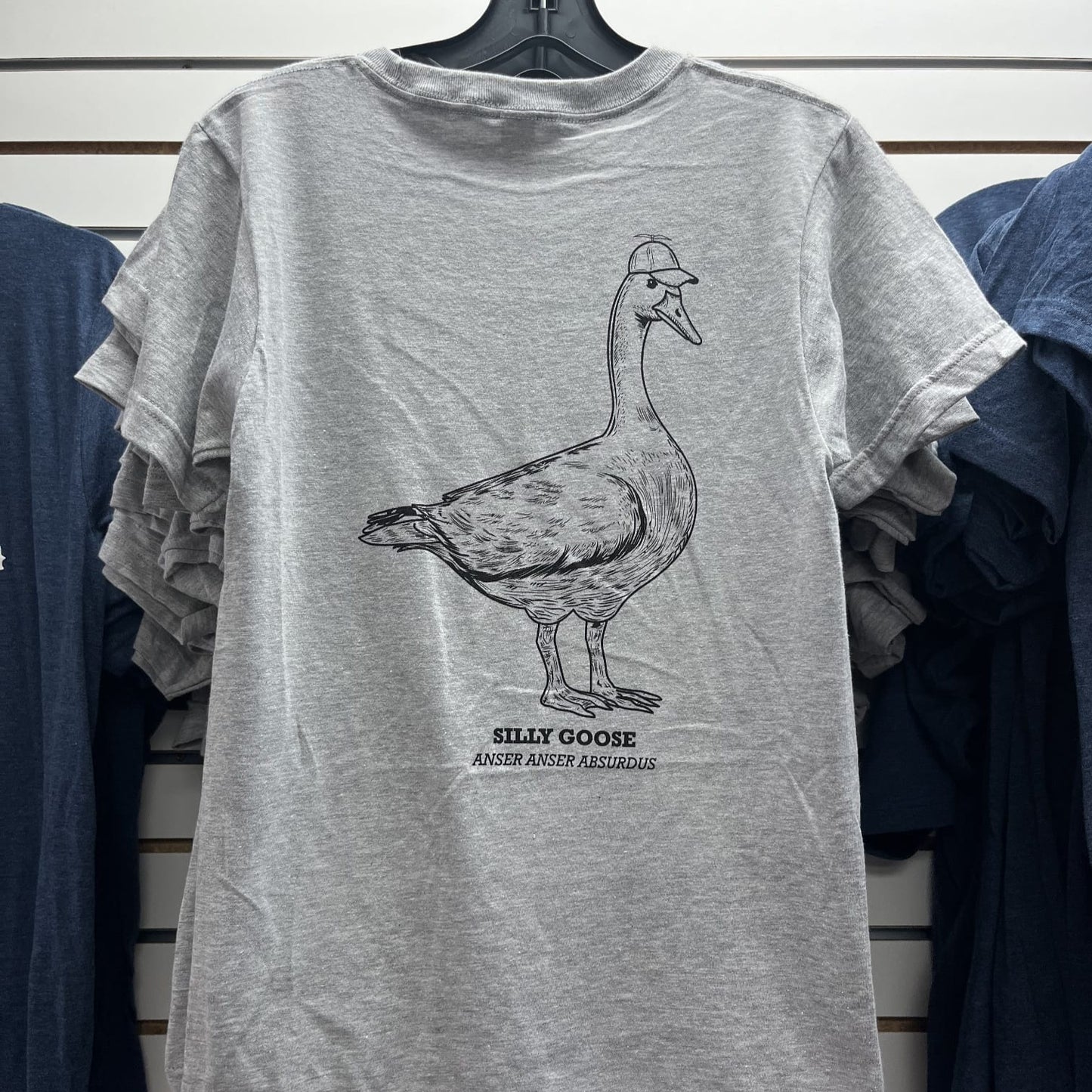 Gray t-shirt with a goose graphic and text on a hanger against a striped wall.