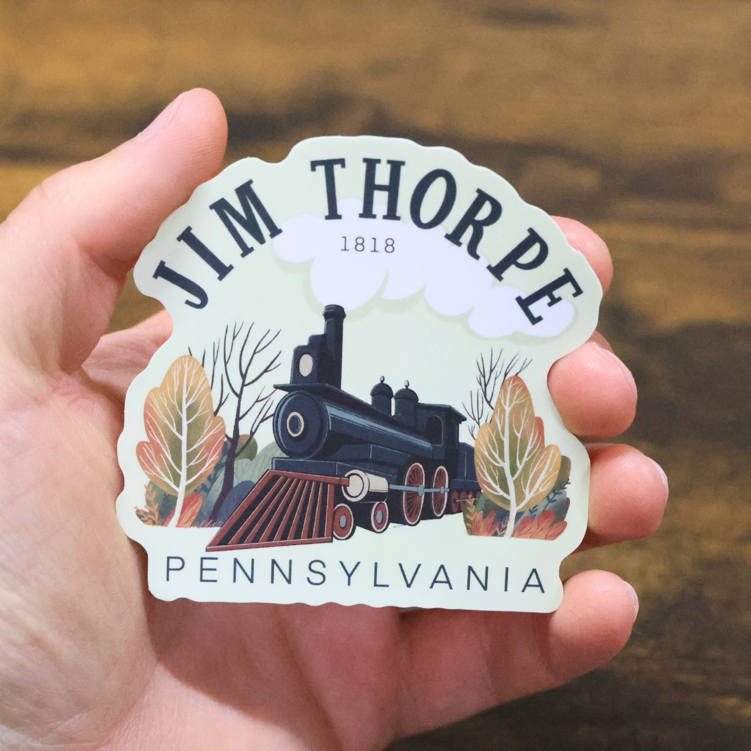 Sticker of Jim Thorpe, Pennsylvania with a train design held in a hand against a wooden background.