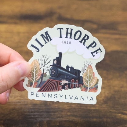 Sticker of a steam train with 'Jim Thorpe, Pennsylvania' text held by a hand on a wooden surface.