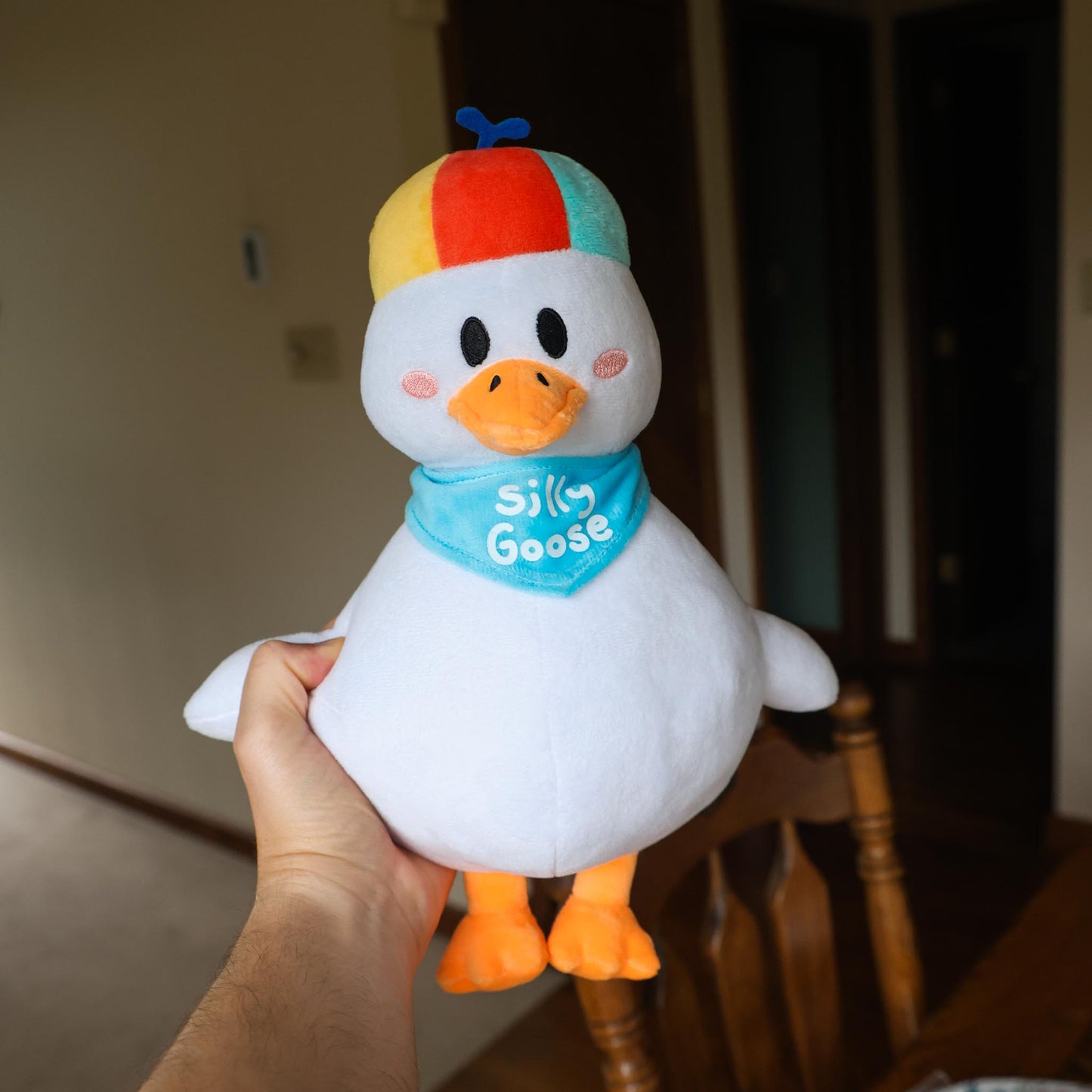 Plush toy goose with colorful hat held by a hand in an indoor setting