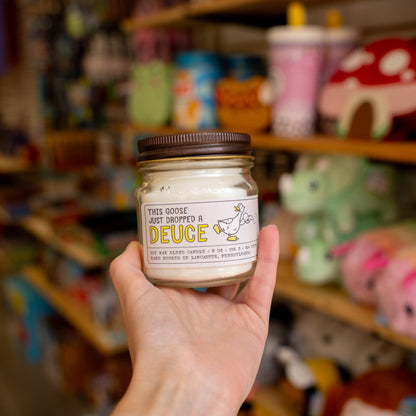 Hand holding a Silly Goose candle jar labeled 'DEUCE' in a store setting with shelves in the background.