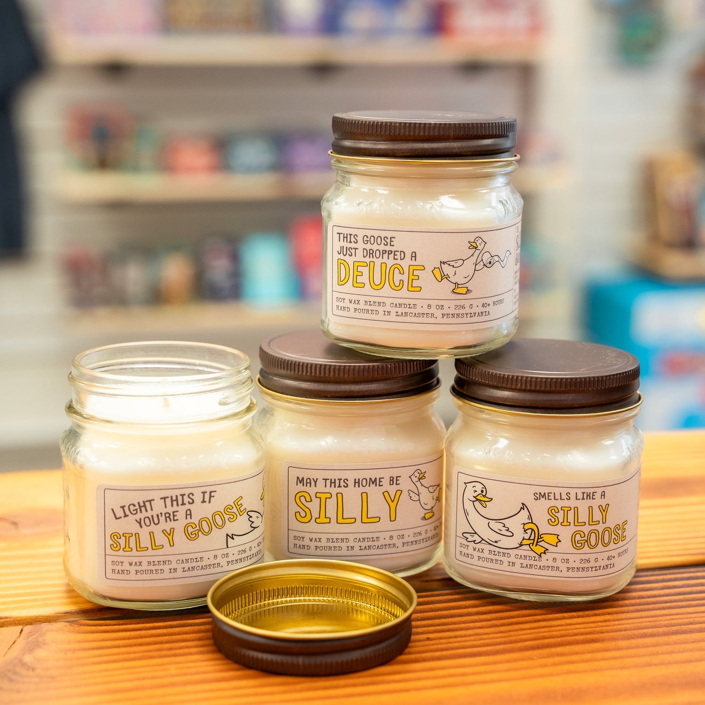 Silly Goose Four jars of candles with humorous labels on a wooden surface.